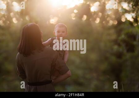 Junge Mutter hält Baby in den Armen in den Strahlen des Sonnenuntergangs im Park, das Mädchen schaut von hinten. Die Frau küsst, umarmt Tochter. Mütterliche Versorgung, Stockfoto