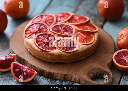 Saftiger hausgemachter Kuchen mit blutroten Orangenscheiben serviert mit rohen Orangen auf Eiche Schneidebrett über schäbig rustikalen blauen Hintergrund Nahaufnahme Stockfoto