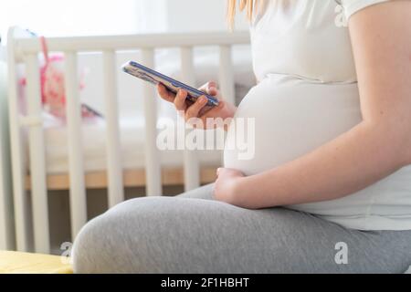 Nahaufnahme des Mittelabschnittes einer unbekannten schwangeren Frau aus Kaukasien in der Hand Handy und Bauch während der Schwangerschaft während des Sitzens Die Wiege in hellen r Stockfoto