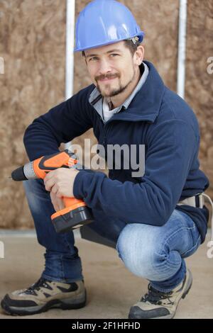 Reparaturman trägt Hardhat lächelnd posiert mit einem Bohrer Stockfoto
