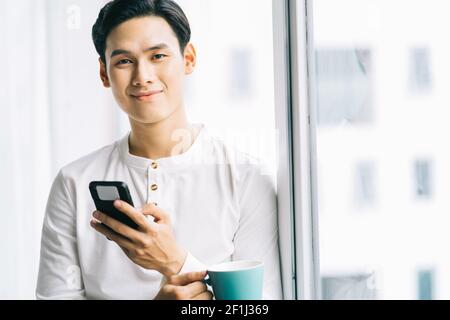 Asiatischer Geschäftsmann trank Kaffee und berührte das Telefon Das Fenster während der Pause Stockfoto