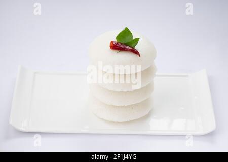 Iddly oder Idli, südindisches Hauptfrühstück garniert mit rotem trockenem Chili und Curryblättern, das wunderschön auf weißem rechteckigem Sockel angeordnet ist Stockfoto