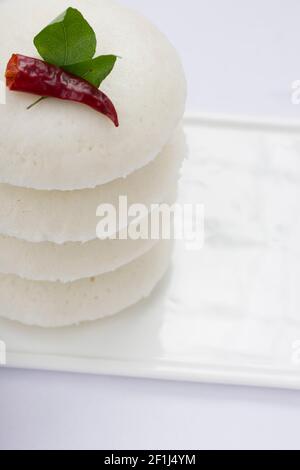 Iddly oder Idli, südindisches Hauptfrühstück garniert mit rotem trockenem Chili und Curryblättern, das wunderschön auf weißem rechteckigem Sockel angeordnet ist Stockfoto