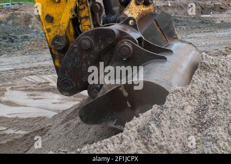 Der Eisenmetalleimer eines großen gelben Baggers gräbt einen Erdhaufen auf einer Baustelle, während er die Straße gegen den Rücken überzieht Stockfoto