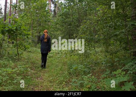 Ein rothaariges Mädchen in einer schwarzen Jacke und Leggings ist Wandern entlang eines Pfades auf dem grünen Gras in einem Waldlichtung zwischen hohen Bäumen mit saftigen Blättern Stockfoto