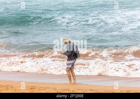 Strandangeln, ältere Männer, die an einem Strand in Sydney in Australien fischen, tragen Shorts und einen Hut Stockfoto