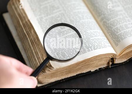 Vintage alten historischen Papier Textbuch auf Tisch geöffnet und Lupe in menschlicher Hand. Bibel, Wörterbuch, antike Dokumentenrecherche mit Lupe, wisd Stockfoto