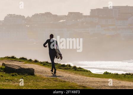 Ein männlicher Surfer, der sein Surfbrett trägt und den Küstenpfad entlang in Richtung Fistral in Newquay in Cornwall läuft. Stockfoto