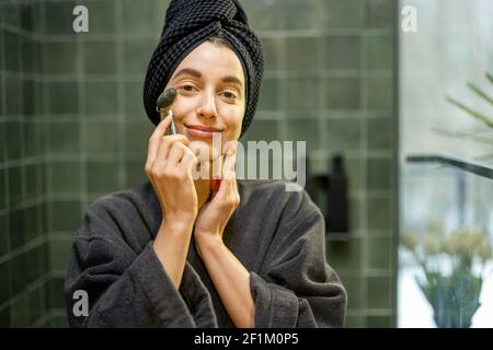 Frau massiert ihr Gesicht mit Roller, in Robe mit Haaren in einem Handtuch nach der Dusche im grünen Badezimmer zu Hause eingewickelt. Lächelnd in die Kamera. Beauty- und Pflegekonzept. Stockfoto