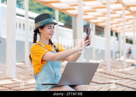 Porträt einer jungen Frau, die per Video-Link am Telefon kommuniziert, auf ihrem Schoß ist ein Laptop. Das Konzept der Freiberufung und Telekommunikation. Stockfoto