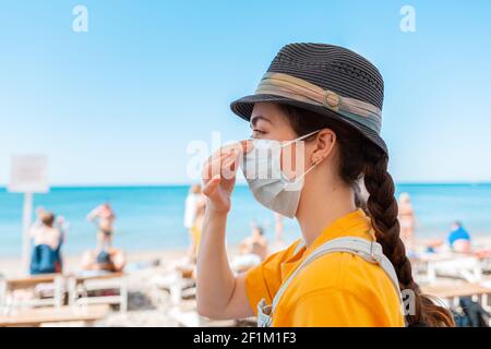 Virenschutz während der Weihnachtszeit. Porträt einer jungen Frau, die eine medizinische Maske auf ihr Gesicht legt. Seitenansicht. Im Hintergrund das Meer und Th Stockfoto