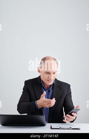 Stirnrunzeln ernst reifen Unternehmer Video Anruf Mitarbeiter über Smartphone, wenn Sitzen am Schreibtisch Stockfoto