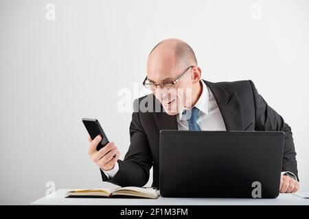 Ein glänzender Geschäftsmann in einer Brille, der am Schreibtisch sitzt und geöffnet ist Laptop und Lesen von Nachrichten über das Scheitern von Geschäftsrivalen Stockfoto