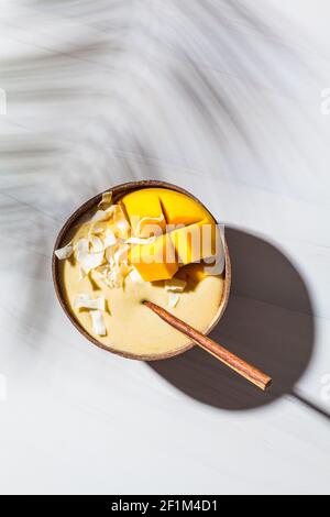 Mango Smoothie Schale mit Kokosflocken in einer Kokosnussschale, weißer Marmorhintergrund, Draufsicht. Veganes Lebensmittelkonzept. Stockfoto