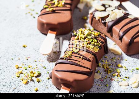 Schokoladeneis Popsicle mit Mandeln, Kokosnuss und Pistazien auf grauem Hintergrund bestreut. Veganes Eis in dunkler Schokolade, Draufsicht. Stockfoto