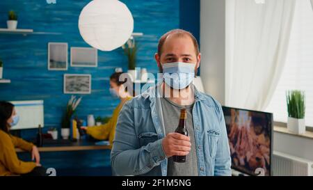 Mann mit Schutzmaske Blick auf Kamera hält Flasche Bier bereit für Party mit Freunden im Wohnzimmer Respekt soziale Distanz in der globalen Pandemie. Menschen sozialisieren während covid 19 Ausbruch Stockfoto