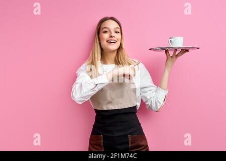 Kellnerin Frau in Schürze hält Tablett mit Tasse Kaffee, Blick mit Überraschung Gesicht zeigt Finger auf Tasse. Isoliert rosa Studio Hintergrund, Porträt Stockfoto