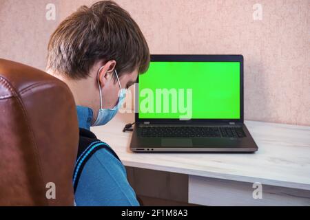 Mann in einer medizinischen Gesichtsmaske schläft am Arbeitsplatz In der Nähe eines Laptops mit grünem Bildschirm Stockfoto