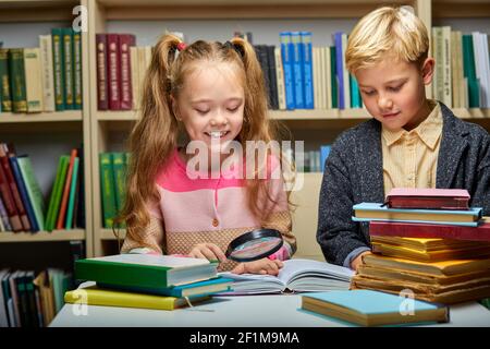 Fleißiger Schüler mit Büchern in der Bibliothek, Junge und Mädchen verwenden Lupe für besseres Lernen und Lesen Stockfoto