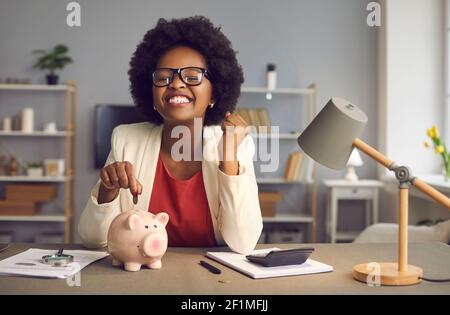 Aufgeregt glücklich afroamerikanische Frau mit ja Geste setzen Münze Im Sparschwein Stockfoto