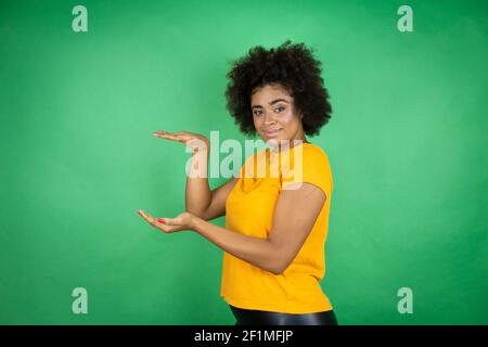 afroamerikanische Frau trägt orange Casual Shirt über grünem Hintergrund gestikulieren mit Händen zeigt große und große Größe Zeichen, Maßnahme Symbol. Lächelnd Stockfoto