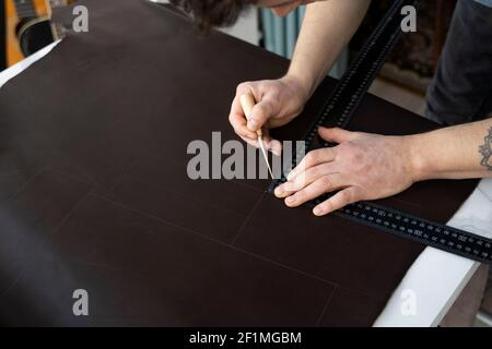 Herrenhand, die eine Ahle und ein Metalllineal hält und an einer Ledertasche in seiner Werkstatt arbeitet. Arbeitsprozess mit einem braunen Naturleder. Handwerker Stockfoto