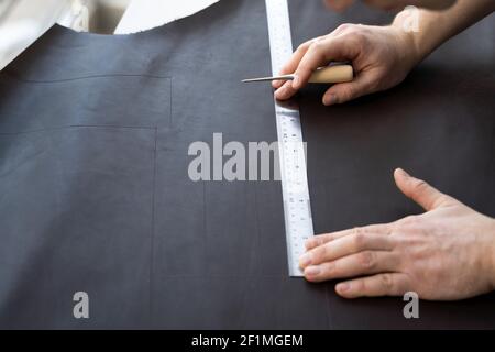 Herrenhand, die eine Ahle und ein Metalllineal hält und an einer Ledertasche in seiner Werkstatt arbeitet. Arbeitsprozess mit einem braunen Naturleder. Handwerker Stockfoto