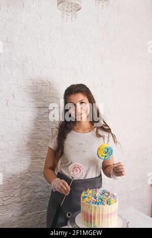 Die Konditorin hält zwei große Lutscher in den Händen und steht neben dem zubereiteten Kuchen. Backwaren auf Bestellung Stockfoto