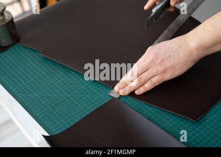 Herrenhand hält ein Schreibwarenmesser und ein Metalllineal und schneidet auf einem Stück für eine Ledertasche in seiner Werkstatt. Arbeitsprozess mit einem braunen Stockfoto