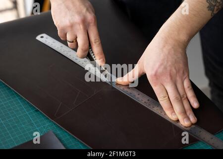 Herrenhand hält ein Schreibwarenmesser und ein Metalllineal und schneidet auf einem Stück für eine Ledertasche in seiner Werkstatt. Arbeitsprozess mit einem braunen Stockfoto