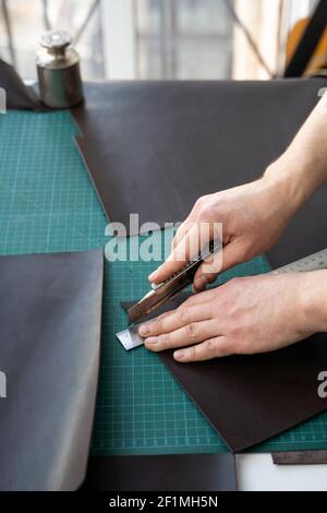 Herrenhand hält ein Schreibwarenmesser und ein Metalllineal und schneidet auf einem Stück für eine Ledertasche in seiner Werkstatt. Arbeitsprozess mit einem braunen Stockfoto