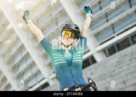 Porträt einer erfolgreichen Radfahrerin in Radbekleidung hob ihre Arme und lächelte der Kamera zu, während sie tagsüber mit ihrem Fahrrad im Freien stand. Aktiver Lebensstil, Sportkonzept Stockfoto