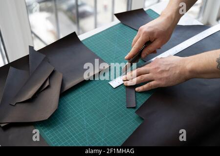 Herrenhand hält ein Schreibwarenmesser und ein Metalllineal und schneidet auf einem Stück für eine Ledertasche in seiner Werkstatt. Arbeitsprozess mit einem braunen Stockfoto