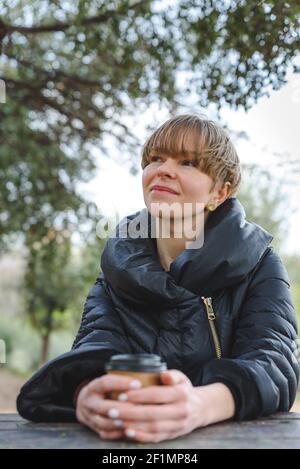 Schöne junge blonde Frau sitzt neben einem Holztisch in einem grünen Park. Sie trägt einen trendigen schwarzen Mantel und hat eine Einweg-Papierkaffeetasse an ihren Händen. Sie hat einen nachdenklichen Blick und Ausdruck. Stockfoto