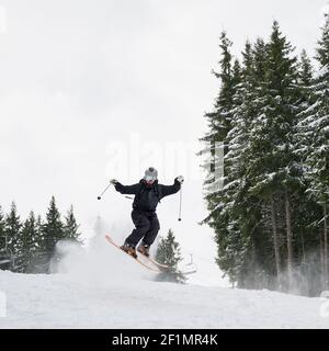 Männliche Skifahrer im Skianzug und Helm Skifahren auf frischem Pulverschnee mit Nadelbäumen und Himmel im Hintergrund. Mann Freerider in Skibrille macht Sprung beim Rutschen auf schneebedeckten Pisten. Stockfoto