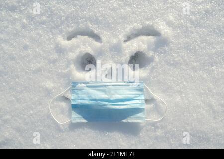 Ein aufgeregter Smiley malte im Schnee mit einer echten medizinischen Maske auf seinem Gesicht. Gesundheitskonzept. Stockfoto