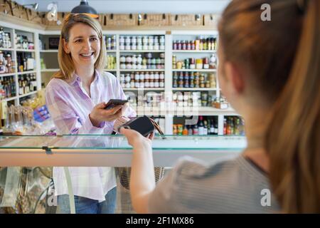 Lächelnd Weiblich Kunde Feinkostgeschäft Herstellung Kontaktlose Zahlung Mit Mobiltelefon Für Shopping Zum Sales Assistant Stockfoto