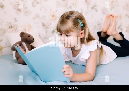 Ein kleines Mädchen liegt auf dem Bett im Schlafzimmer und liest ein blaues Buch. Bildungskonzept. Schulbildung zu Hause. Hausaufgaben. Hochwertige Fotos Stockfoto