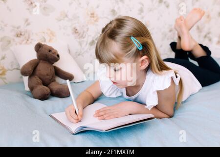 Ein kleines Mädchen liegt auf dem Bett im Schlafzimmer und schreibt in einem blauen Buch. Bildungskonzept. Schulbildung zu Hause. Hausaufgaben. Hochwertige Fotos Stockfoto