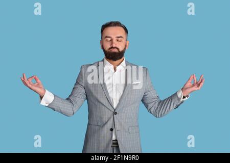 Konzept für das Stressmanagement am Arbeitsplatz. Friedlicher Büroangestellter meditiert mit geschlossenen Augen auf blauem Studiohintergrund. Junger Geschäftsmann fühlt sich ruhig an Stockfoto