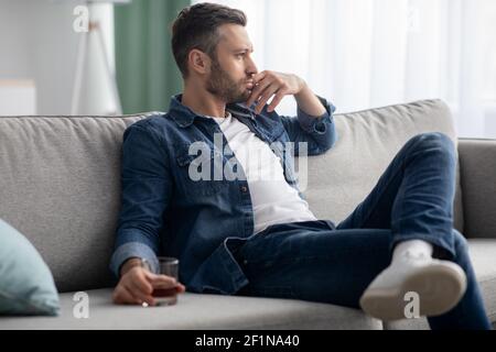 Depression, Krisenkonzept. Trauriger bärtiger Mann mittleren Alters, der auf dem Sofa sitzt, zu Hause Alkohol trinkt, sich einsam und krank fühlt, ein Glas Whiskey A hält Stockfoto