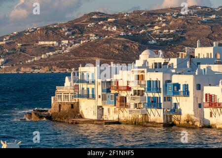 Blick auf Little Venice auf mykonos mit bunten Fenstern und Balkonen in Blau und Rot. Im Hintergrund ein Hügel mit winzigen Häusern, alle vom Meer umgeben Stockfoto