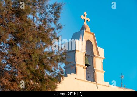 Ein Baum und ein weißes Marmorkreuz auf der Spitze Ein Turm mit Bronzeglocke vor blauem Himmel Auf einer griechischen Insel Stockfoto