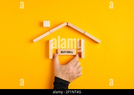 Männliche Hand, die ein Kauf- oder Verkaufsschild in einem Haus aus Holzblöcken auf gelbem Hintergrund platziert. Kauf oder Verkauf einer Schlauch-Entscheidung in der Immobilienmarke Stockfoto