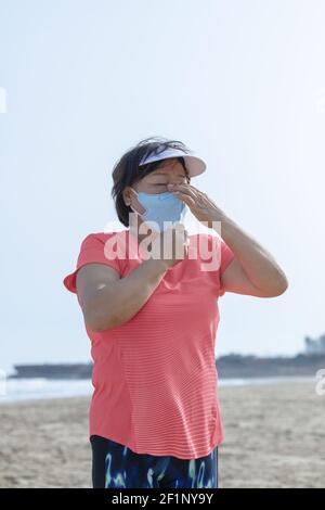 Reife asiatische Frau trägt ein Visier Fixierung ihrer Gesichtsmaske am Strand auf einem unscharf Hintergrund. Selektiver Fokus und Nahaufnahme. Übungskonzept. Stockfoto