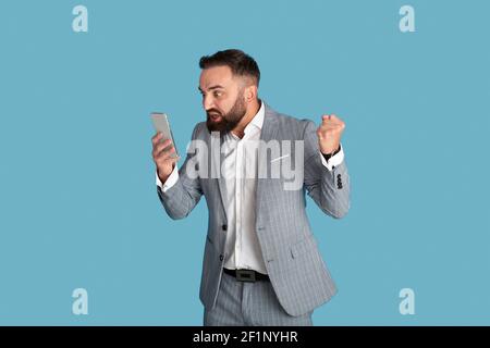 Aggressiver junger Geschäftsmann, der unter Stress am Arbeitsplatz leidet und das Smartphone über blauem Studiohintergrund anruft. Wütender Mitarbeiter im Büro, tausendjährig Stockfoto