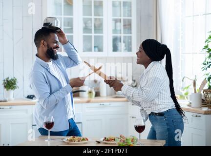 Fröhliche schwarze Paar Spaß in der Küche, spielerisch kämpfen mit Geschirr, zielte sich mit Utensilien Werkzeuge und lachen, junge afrikanische Spou Stockfoto