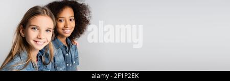 Zwei glückliche interracial Mädchen in Denim-Kleidung lächeln an Kamera isoliert auf grau, Banner Stockfoto