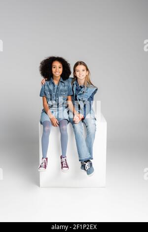 Modische interracial Kinder in Denim-Kleidung auf weißen Würfel sitzen Auf Grau Stockfoto