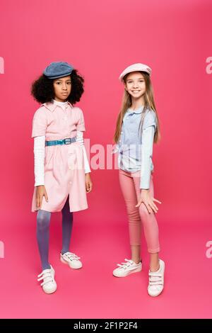 Interracial Kinder in trendigen Kleidung, Turnschuhe und Mützen Blick auf Kamera, während posiert auf rosa Stockfoto